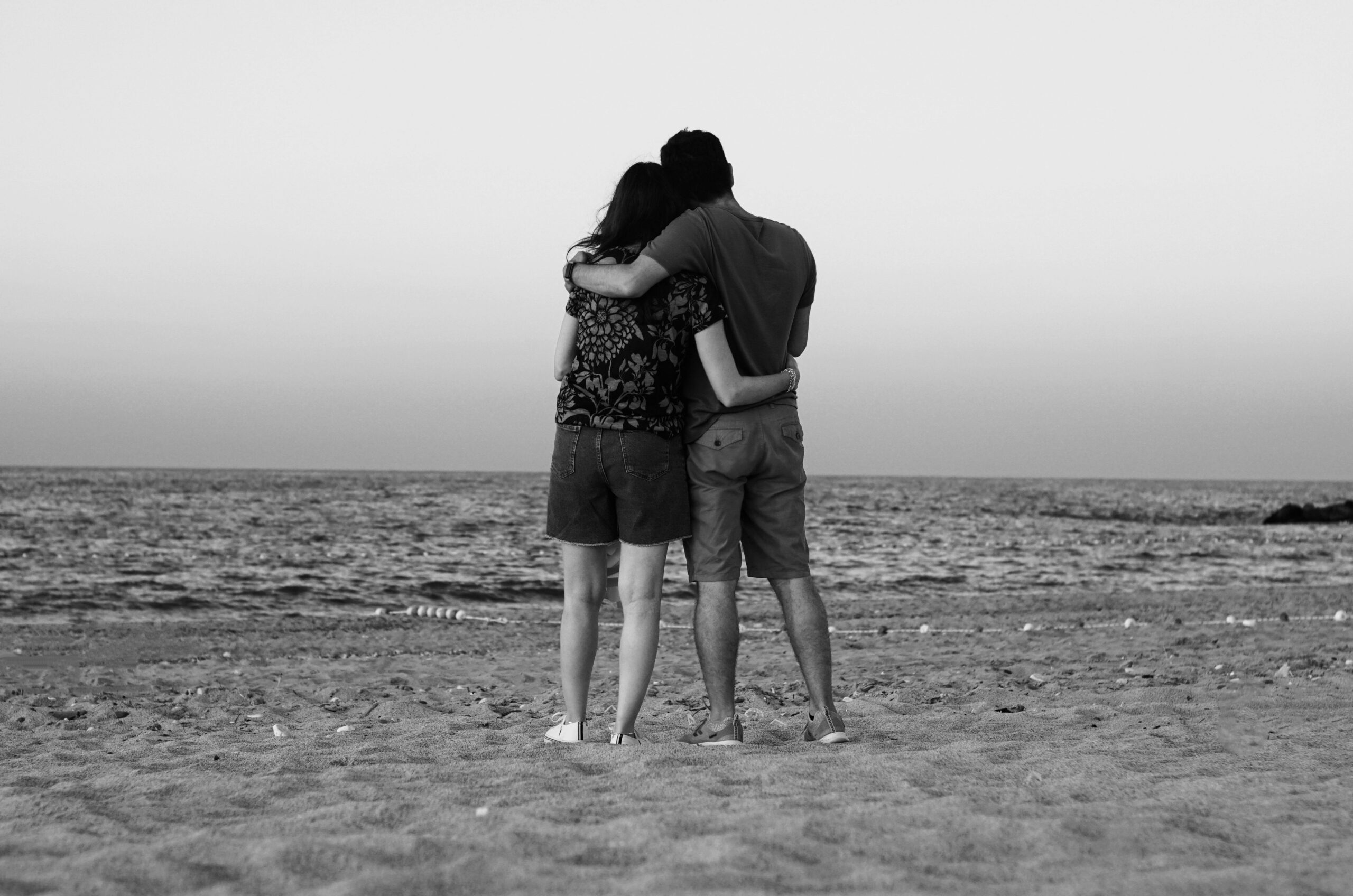 A loving couple embraces while standing on a tranquil beach, captured in black and white.
